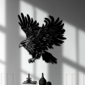 Może przedstawiać: Czarna metalowa dekoracja ścienna w kształcie orła z detalicznym wzorem. Orzeł znajduje się w pozycji lotu z rozpostartymi skrzydłami. Dekoracja jest zamontowana na białej ścianie nad czarną konsolą z dwoma szufladami.