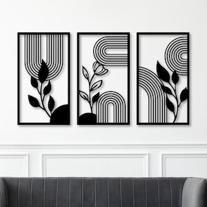 以下が含まれることがあります： 白を背景にした黒いフレームの壁掛けアート3点。それぞれ、黒いアーチ、葉、花の要素の異なるデザインが特徴です。アートはグレーのソファの上に展示されています。