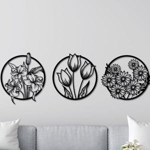 Könnte beinhalten: Drei Wandkunststücke mit schwarzen Metallblumen. Jedes kreisförmige Design zeigt eine andere Blume: Lilien, Tulpen und Gänseblümchen. Die Kunst ist an einer weißen Wand über einem grauen Sofa angebracht.