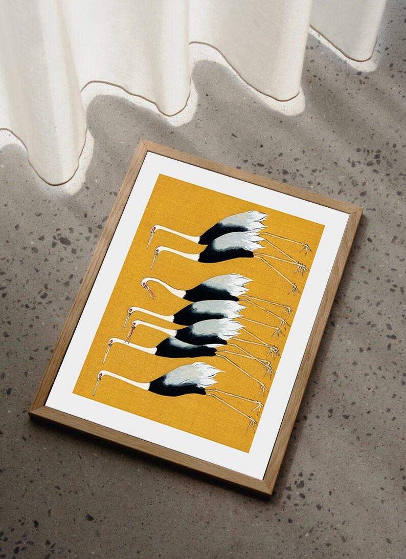 May include: A framed print featuring a flock of cranes in flight. The birds have white and black plumage with long, thin legs and beaks. The background is a solid yellow, and the frame is made of light-colored wood.