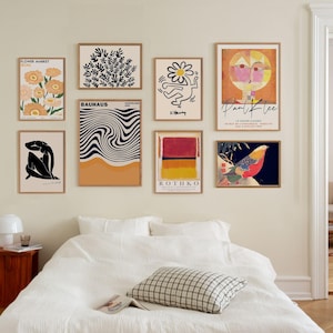 May include: A bedroom interior with a white bed and a wall gallery of nine framed prints. The prints feature abstract art, floral designs, and a bird illustration. The prints are arranged in a grid pattern on the wall.