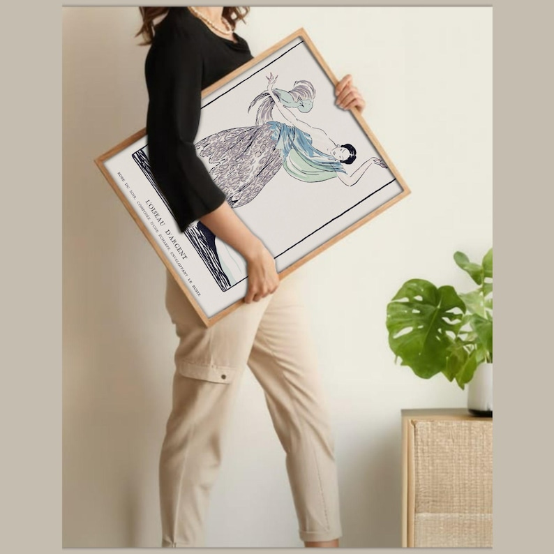 May include: A framed art print featuring a stylized illustration of a woman in flowing attire. The artwork, in shades of blue, gray, and black, is set against a white background. The frame is a light wood color, and the print includes the text "L'OISEAU D'ARGENT".