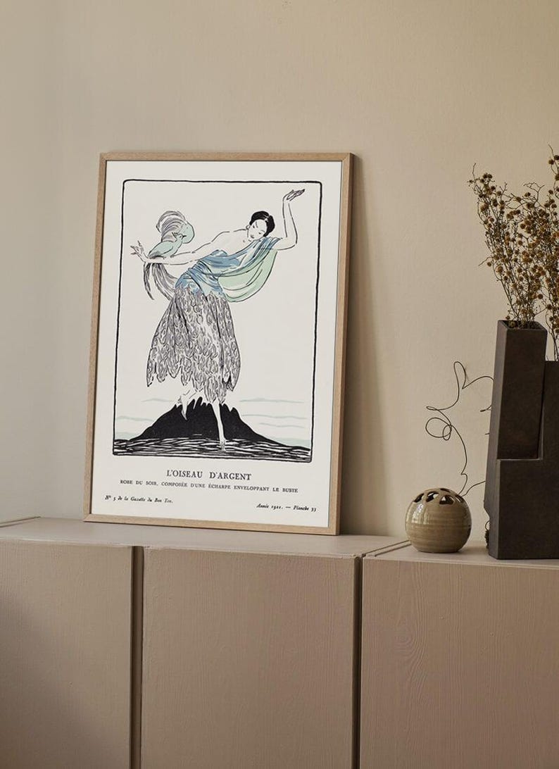 May include: Framed art print featuring a stylized illustration of a woman in a flowing dress, with a blue and green scarf. The artwork is in black and white with a mountain-like shape at the bottom. The print is titled "L'OISEAU D'ARGENT".