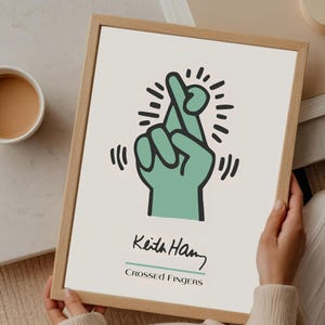 May include: A framed art print featuring a stylized green hand with crossed fingers, surrounded by radiating lines. The print includes the artist's signature, "Keith Haring," and the text "Crossed Fingers" in black font.