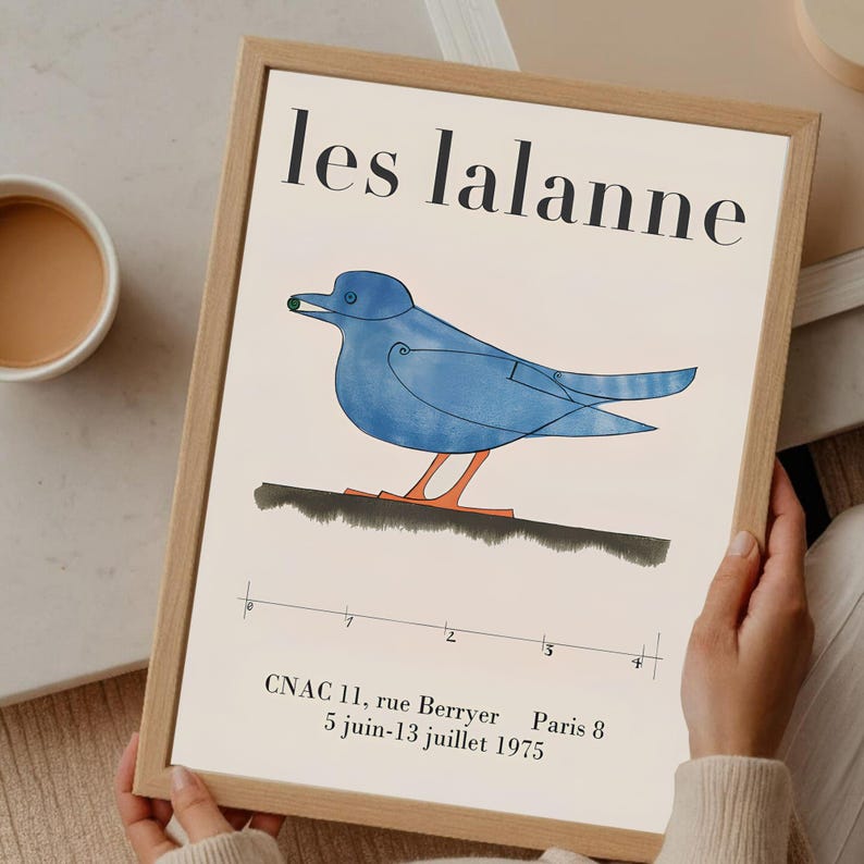 May include: A framed art print featuring a blue bird illustration with orange legs and a green dot in its beak. The print has the text "les lalanne" at the top and additional text at the bottom, all within a light wood frame.
