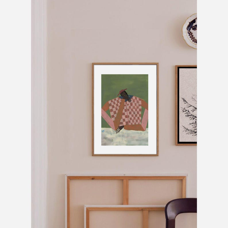 May include: A framed art print featuring a figure in a checkered shirt and a red hat, set against a green backdrop. The artwork has a modern, abstract style with a focus on color and form. The frame is a warm brown.