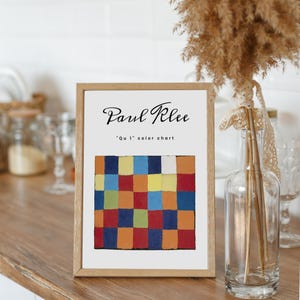 May include: A framed print of a Paul Klee artwork titled "Qu 1 color chart." The artwork features a grid of colorful squares in shades of red, orange, blue, yellow, and green. The frame is made of light-colored wood.