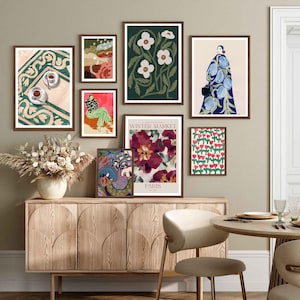 May include: A collection of framed art prints featuring various designs, including floral patterns, abstract shapes, and a woman in a blue dress. The prints are arranged on a wall above a wooden cabinet and a dining table with chairs.