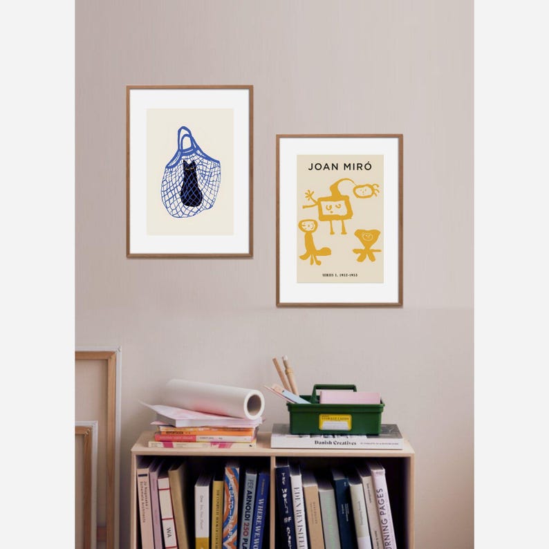 May include: Two framed art prints. One features a black cat in a blue net bag on a cream background. The other print displays abstract yellow figures with the text "JOAN MIRO" and dates. Both are in brown wooden frames.