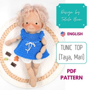 May include: A crocheted doll wearing a blue tunic top and white shorts. The doll has light brown skin, blonde hair, and is sitting on a round, woven mat. The image includes text that says "Design by Talula Boom", "ENGLISH", and "TUNIC TOP (Taya, Mari) PDF PATTERN".