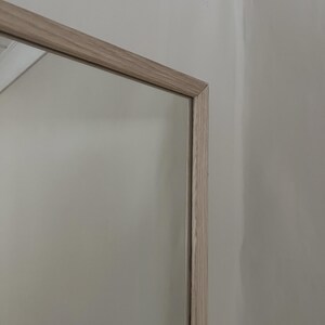 May include: A rectangular mirror with a light-colored wooden frame. The frame has a natural wood grain appearance. The mirror reflects a neutral-colored wall. The frame's corner is visible.
