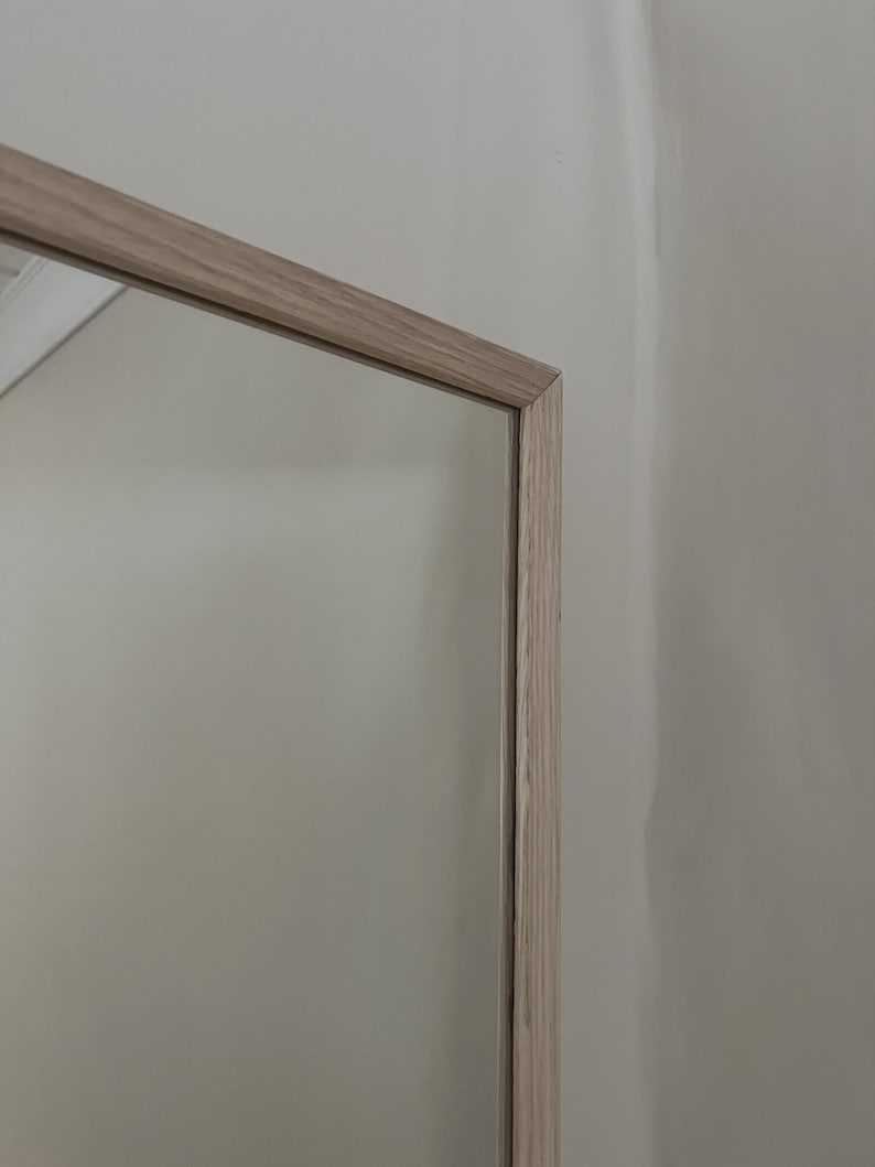 May include: A rectangular mirror with a light-colored wooden frame. The frame has a natural wood grain appearance. The mirror reflects a neutral-colored wall. The frame's corner is visible.