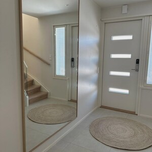 May include: A large rectangular mirror with a light wood frame is mounted on a white wall. A round jute rug sits on the tiled floor. The mirror reflects a staircase and another round rug.