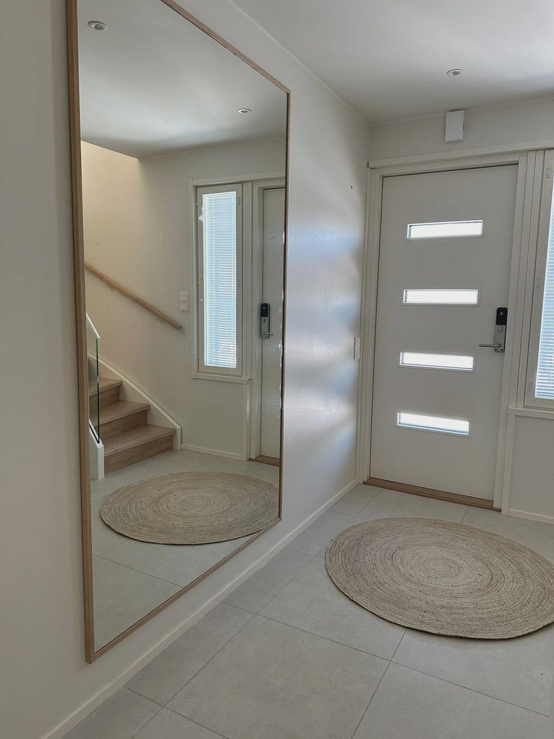 May include: A large rectangular mirror with a light wood frame is mounted on a white wall. A round jute rug sits on the tiled floor. The mirror reflects a staircase and another round rug.