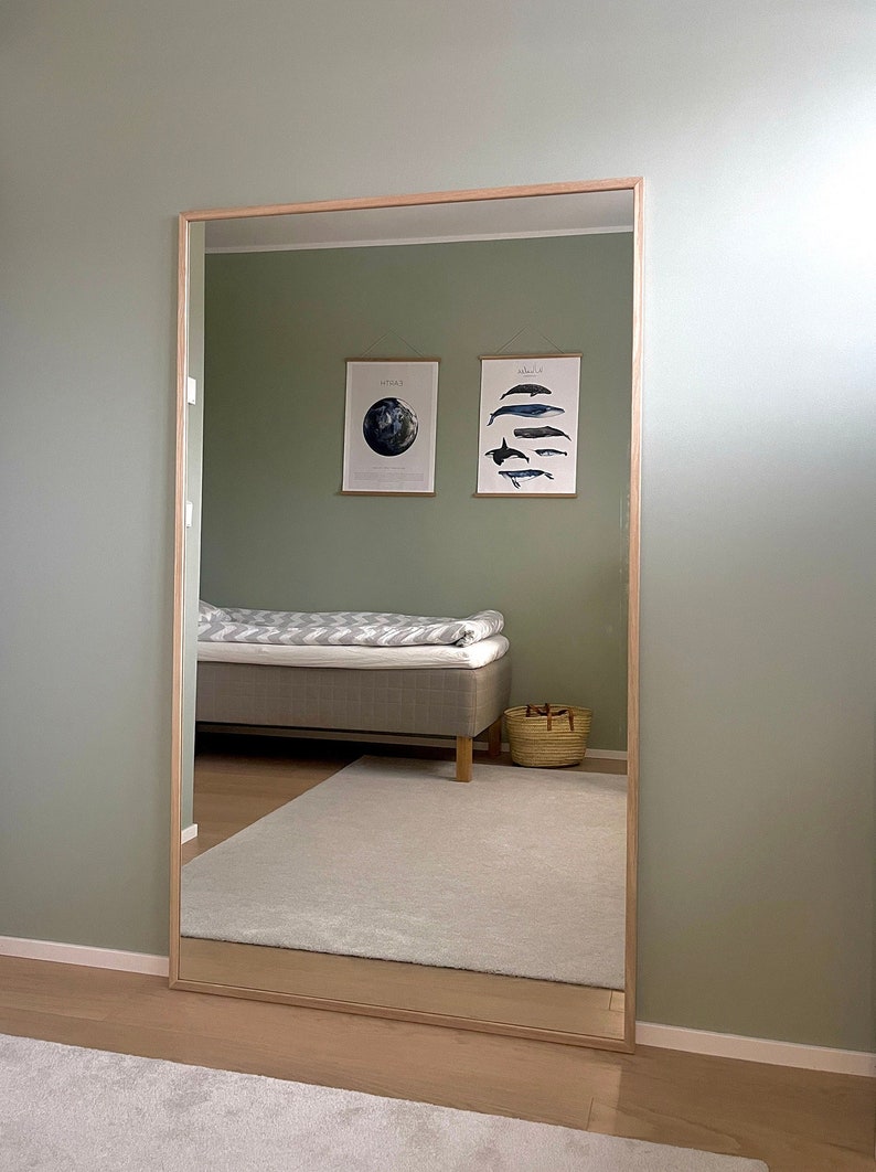 May include: A large rectangular mirror with a light wood frame. The mirror reflects a bedroom with a bed, two framed prints, and a woven basket. The walls are painted a muted green, and the floor is light wood.