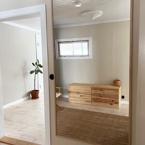 May include: A large rectangular mirror with a light wood frame is mounted on a beige wall. The mirror reflects a room with a wooden dresser, a woven rug, and a window. The room has light-colored walls and a white ceiling.