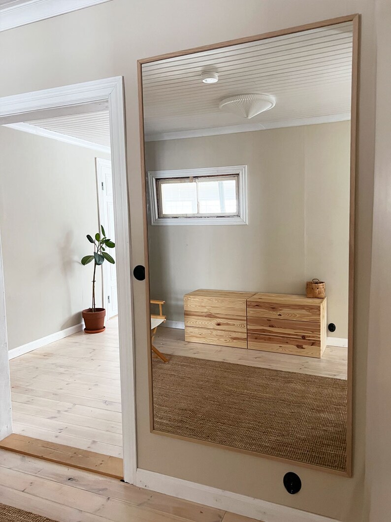 May include: A large rectangular mirror with a light wood frame is mounted on a beige wall. The mirror reflects a room with a wooden dresser, a woven rug, and a window. The room has light-colored walls and a white ceiling.