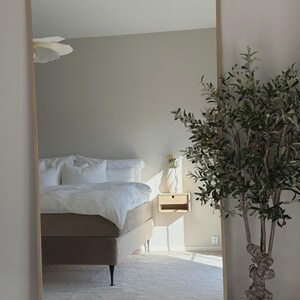 May include: A large, rectangular, full-length mirror with a light wood frame. The mirror reflects a bedroom scene with a bed, nightstand, and a potted olive tree. The room has neutral tones and a minimalist aesthetic.