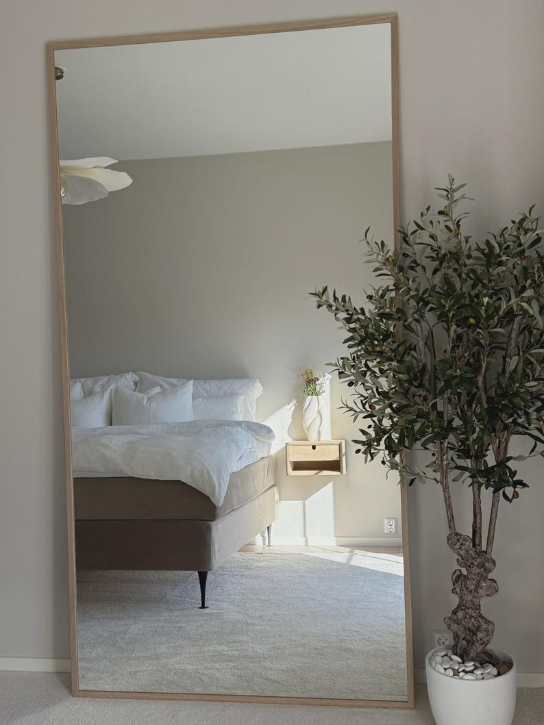 May include: A large, rectangular, full-length mirror with a light wood frame. The mirror reflects a bedroom scene with a bed, nightstand, and a potted olive tree. The room has neutral tones and a minimalist aesthetic.