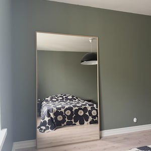 May include: A full-length rectangular mirror with a light wood frame. The mirror reflects a bedroom with a bed covered in a black and white floral patterned quilt. The walls are painted a muted green.
