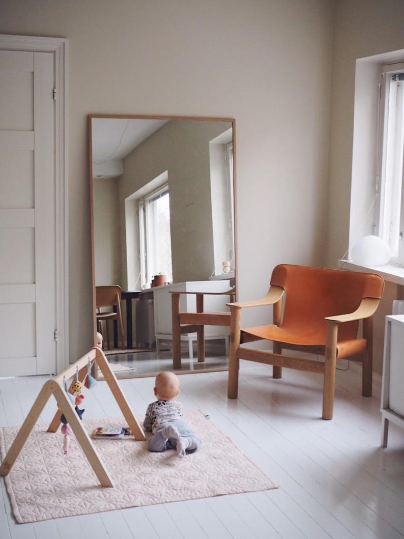 May include: A light-filled room with a large wooden-framed mirror, an orange leather armchair, and a wooden baby gym. A baby is on a pink quilted play mat. The room has white painted wood floors and a neutral wall color.
