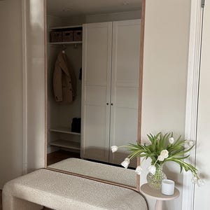 May include: A large rectangular mirror with a wooden frame is mounted on a wall. Below the mirror is a cream-colored, curved bench. A small, light pink side table holds a vase of white tulips and a small white candle.