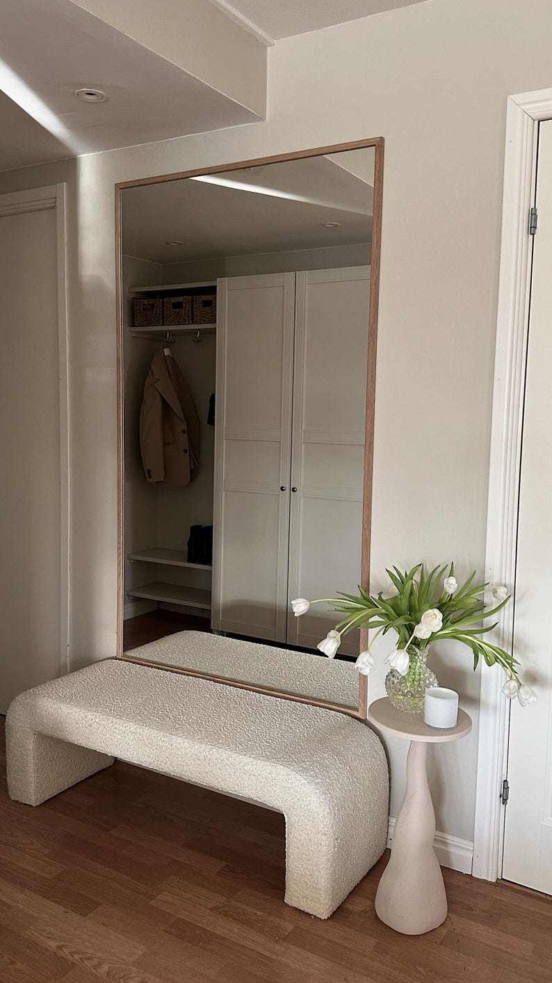 May include: A large rectangular mirror with a wooden frame is mounted on a wall. Below the mirror is a cream-colored, curved bench. A small, light pink side table holds a vase of white tulips and a small white candle.