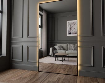 May include: A full-length mirror with a black frame and warm white LED lights around the perimeter. The mirror is reflecting a living room with a couch, coffee table, and a framed print on the wall.