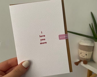 Puede incluir: Una tarjeta de felicitación blanca con una superficie texturizada que presenta el texto rojo i love you more centrado en el frente. La tarjeta se combina con un sobre marrón y tiene una pequeña pegatina morada con el texto jenna catriens studios.