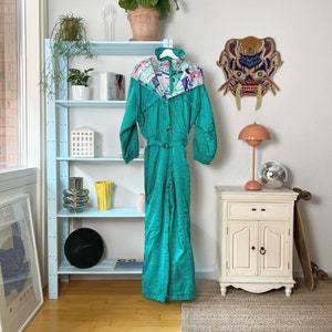 Puede incluir: Un traje de esquí vintage de color turquesa con un colorido estampado geométrico en los hombros cuelga de una percha blanca. El traje de una pieza tiene cintura con cinturón y mangas largas. Una estantería azul claro con plantas y decoración está en el fondo.