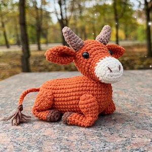May include: A crocheted orange cow figurine with a white muzzle, brown horns, and dark eyes. The handmade plush toy is lying down, with its tail curled. The cow is sitting on a stone surface, with a blurred background of trees.