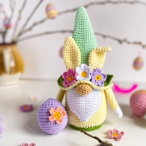 May include: A crocheted Easter gnome with a green hat, yellow ears, and a white beard. The gnome is wearing a flower crown and holding a purple crocheted egg with a flower on it.
