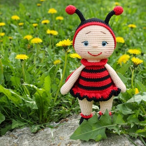 Peut inclure: Une poupée en crochet en forme de coccinelle avec des rayures noires et rouges, des antennes noires et un visage souriant. La poupée est debout dans un champ de pissenlits jaunes.