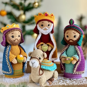 May include: Three crocheted figures representing the Three Wise Men, each wearing a crown and colourful robes, holding small gifts. A small, light brown crocheted camel sits in the foreground. The figures feature detailed stitching and bead accents.
