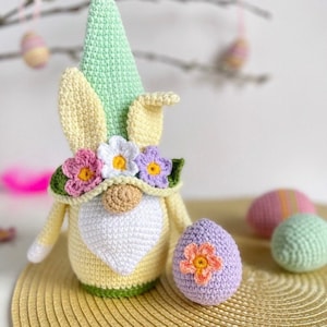 May include: A crocheted Easter gnome with a green hat, yellow body, and white beard. The gnome is wearing a flower crown and holding a purple crocheted egg with a pink flower.