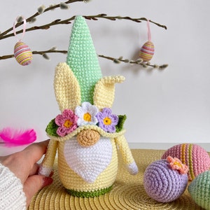 May include: A crocheted Easter gnome with a green hat, yellow and white body, and a flower crown. The gnome is surrounded by crocheted Easter eggs in various colors.