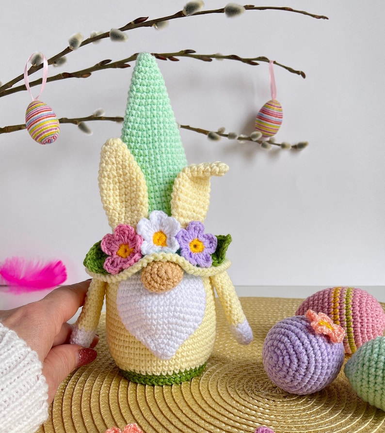 May include: A crocheted Easter gnome with a green hat, yellow and white body, and a flower crown. The gnome is surrounded by crocheted Easter eggs in various colors.