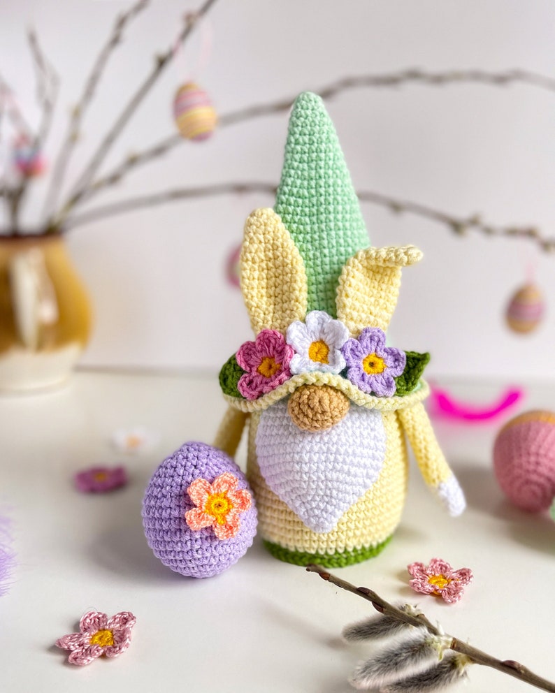 May include: A crocheted Easter gnome with a green hat, yellow ears, and a white beard. The gnome is wearing a flower crown and holding a purple crocheted egg with a flower on it.