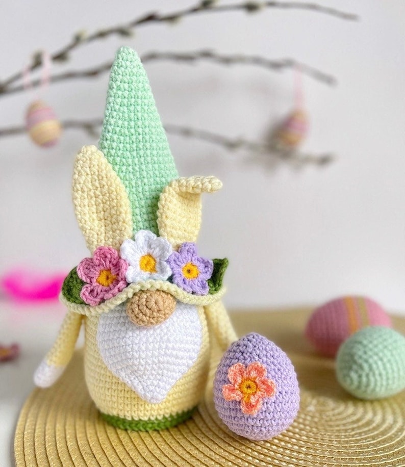 May include: A crocheted Easter gnome with a green hat, yellow body, and white beard. The gnome is wearing a flower crown and holding a purple crocheted egg with a pink flower.