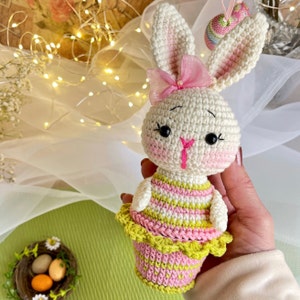 May include: A crocheted bunny rabbit toy wearing a pink and green striped dress with a pink bow. The bunny is holding a basket of pink and green Easter eggs.