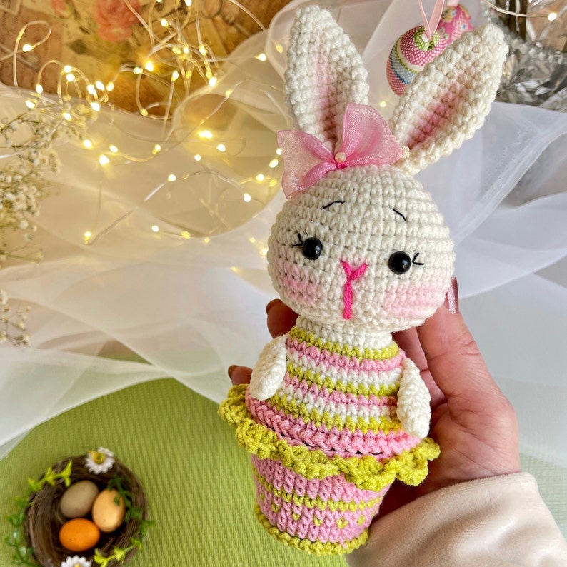 May include: A crocheted bunny rabbit toy wearing a pink and green striped dress with a pink bow. The bunny is holding a basket of pink and green Easter eggs.