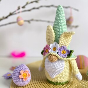 May include: A crocheted Easter gnome with a green hat, yellow and white ears, and a white beard. The gnome is wearing a flower crown with pink, white, and purple flowers. The gnome is sitting on a straw-like placemat with a purple crocheted egg next to it.
