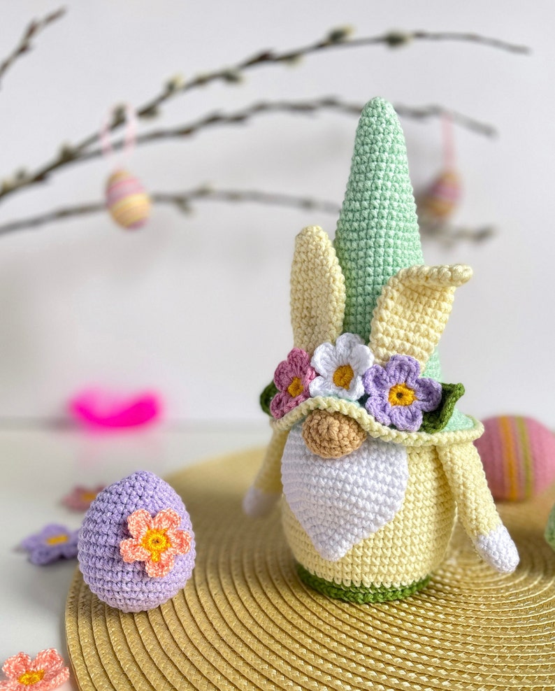 May include: A crocheted Easter gnome with a green hat, yellow and white ears, and a white beard. The gnome is wearing a flower crown with pink, white, and purple flowers. The gnome is sitting on a straw-like placemat with a purple crocheted egg next to it.