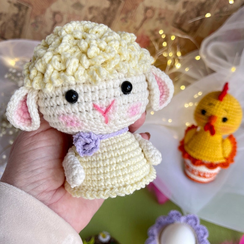 May include: A crocheted white lamb with pink ears and a purple flower on its neck. The lamb is holding a small crocheted egg in its mouth.