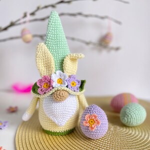 May include: A crocheted Easter gnome with a green hat, yellow bunny ears, and a flower crown. The gnome is holding a purple crocheted Easter egg. There are other crocheted Easter eggs in the background.