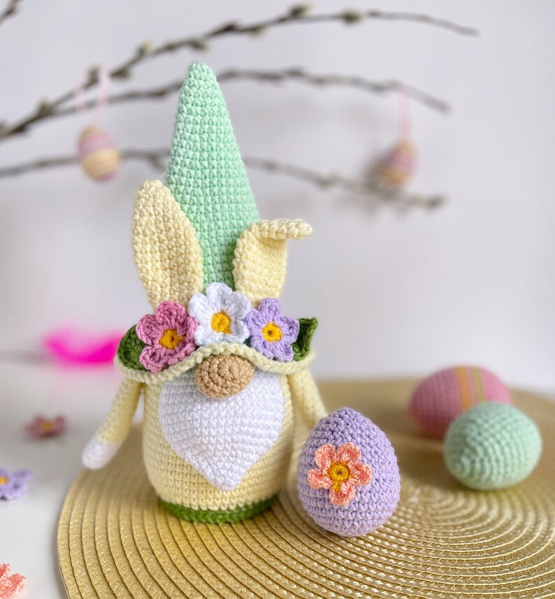 May include: A crocheted Easter gnome with a green hat, yellow bunny ears, and a flower crown. The gnome is holding a purple crocheted Easter egg. There are other crocheted Easter eggs in the background.
