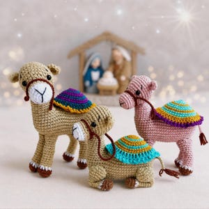 May include: Three crocheted camels in tan and pink with colourful saddle blankets. Each camel has a brown bridle and a small tail. A nativity scene is visible in the background, suggesting a festive theme.