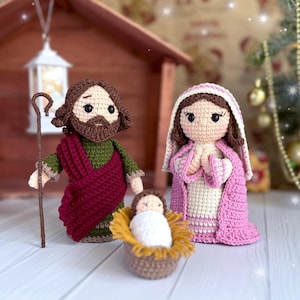May include: Crocheted nativity scene featuring Joseph with a staff, Mary in a pink shawl, and baby Jesus in a manger. The figures are handmade with detailed stitching and soft textures. The scene is set against a backdrop of a wooden structure and a Christmas tree.