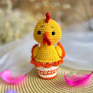 May include: A crocheted yellow chick toy with red and orange accents. The chick is sitting on a woven straw mat.