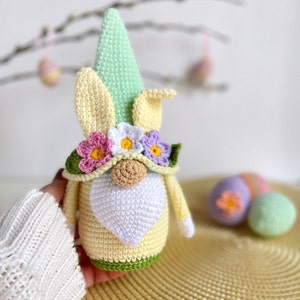 May include: A crocheted Easter gnome with a green hat, yellow body, white beard, and a flower crown. The gnome is holding a green and yellow Easter egg.